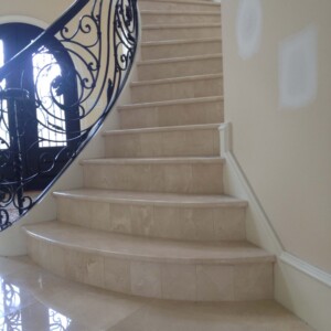 Curved beige marble staircase with ornate black wrought iron railing.