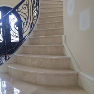 Curved beige marble staircase with ornate black metal railing.