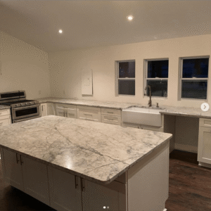 Modern kitchen with marble countertops and farmhouse sink under windows.
