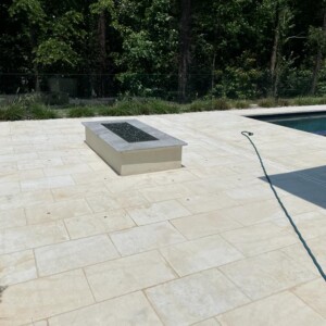Modern outdoor patio with beige tiles, a fire pit, and a pool beside a wooded area.