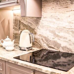 beautiful closeup of custom designed kitchen, with marble looking quartz countertop and backsplash. cream electric kettle with porcelain tea accessories on the marble countertop next to ceramic hob Modern kitchen countertop with electric kettle and teapot set.