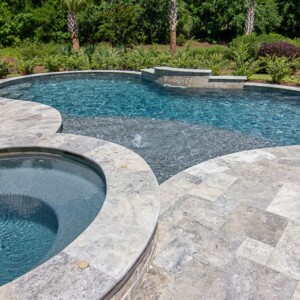 Silver Travertine Pool Deck Pavers San Jose A backyard pool and hot tub with stone tile surrounding and lush greenery.