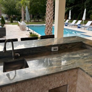 Outdoor kitchen counter with sink overlooking a pool and patio area.